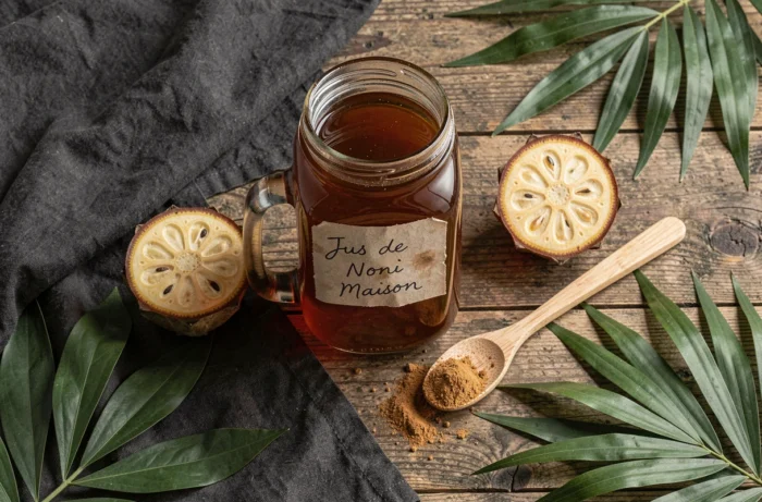 Pot en verre de jus de noni maison fait maison posé sur une table en bois avec des fruits noni frais et feuilles tropicales